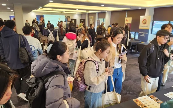 Crowded indoor careers fair with students carrying tote bags and drinks browsing exhibitor stands and banners